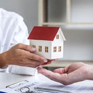 Two people exchanging model house and documents on a table, representing a real estate or housing transaction.